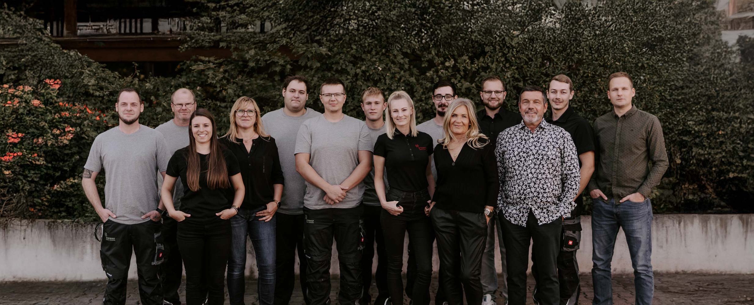 Die Gruppe besteht aus Männern und Frauen unterschiedlichen Alters, die in lässiger Kleidung vor einem bewachsenen Hintergrund stehen.