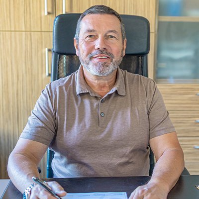 In einem modernen Büro lächelt ein Mann in einem braunen Poloshirt, während er an einem Tisch mit Unterlagen arbeitet.