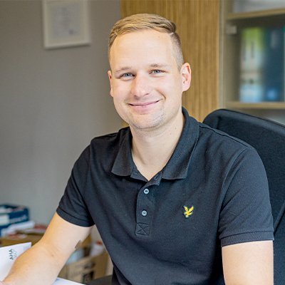 Der Mann trägt ein schwarzes Polohemd und wirkt entspannt, während er in die Kamera lächelt, mit einem hellen Hintergrund.