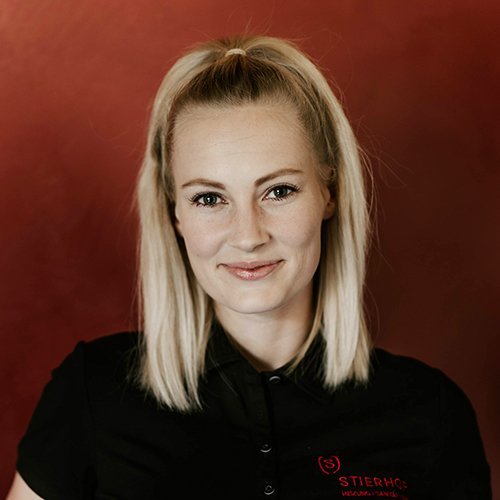 Die Frau mit langen, blonden Haaren und einem Zopf lächelt selbstbewusst in die Kamera, während sie ein schwarzes Oberteil trägt.