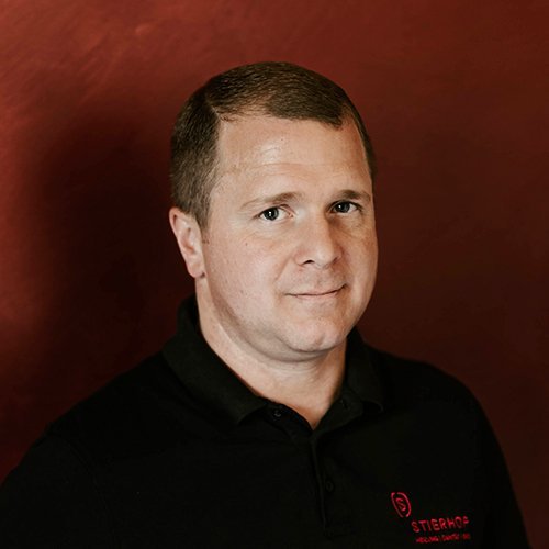 Ein Mann mit kurzem, braunem Haar trägt ein schwarzes Polohemd und schaut direkt in die Kamera vor einem roten Hintergrund.