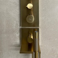 Eine elegante goldene Armatur mit zwei Drehknöpfen und einem Handbrausekopf an einer schlichten Wand aus Beton.