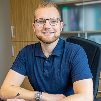 Der Mann trägt ein dunkelblaues Polohemd und hat einen Stift in der Hand, während er an einem Schreibtisch sitzt.