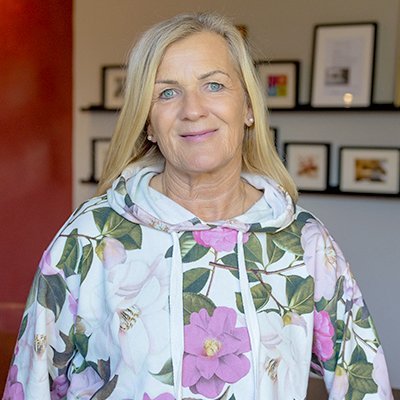 Die Frau in einem blumigen Sweatshirt strahlt Freundlichkeit aus, während sie in einem hellen Raum mit Kunstwerken posiert.
