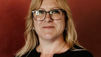 Die Porträtaufnahme zeigt eine selbstbewusste Frau mit langen, blonden Haaren und einer eleganten Brille, die freundlich blickt.