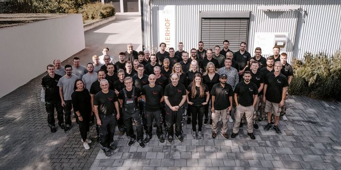 Eine große Gruppe von Mitarbeitern steht lächelnd vor einem modernen Gebäude, gekleidet in schwarze T-Shirts und Arbeitskleidung.